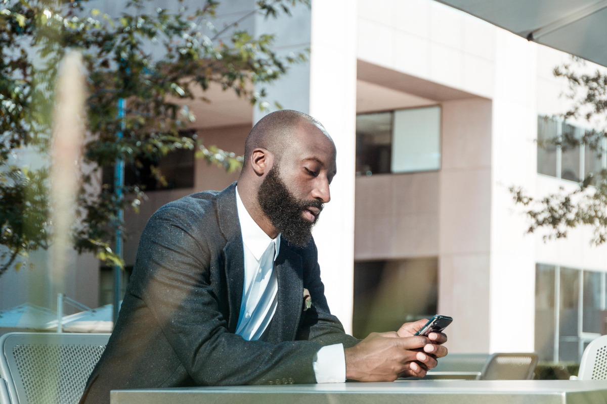 capital gains: Black man in a suit sitting in an outdoor courtyard with a black suit, blue tie, looking at his phone